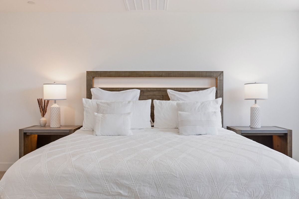 Una cama bien hecha con cubrecolcha blanco acolchado, dos mesitas de noche con lámparas y un cabecero de madera.