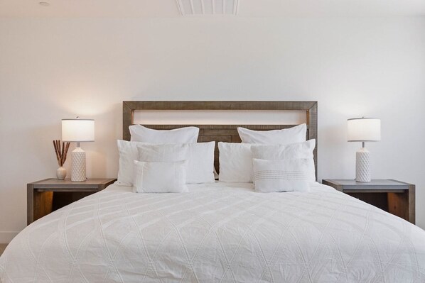 A neatly made bed with a quilted white cover, two bedside tables with lamps, and a wooden headboard. - Luxe Serenity: EV Charger, Pool, Spa & FirePit (Indio)