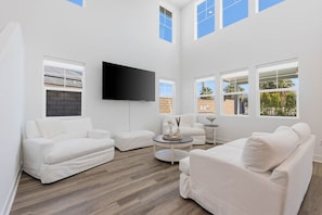 A spacious living room with white sofas, a flat-screen TV, and large windows offering views of the outside. - Luxe Serenity: EV Charger, Pool, Spa & FirePit (Indio)