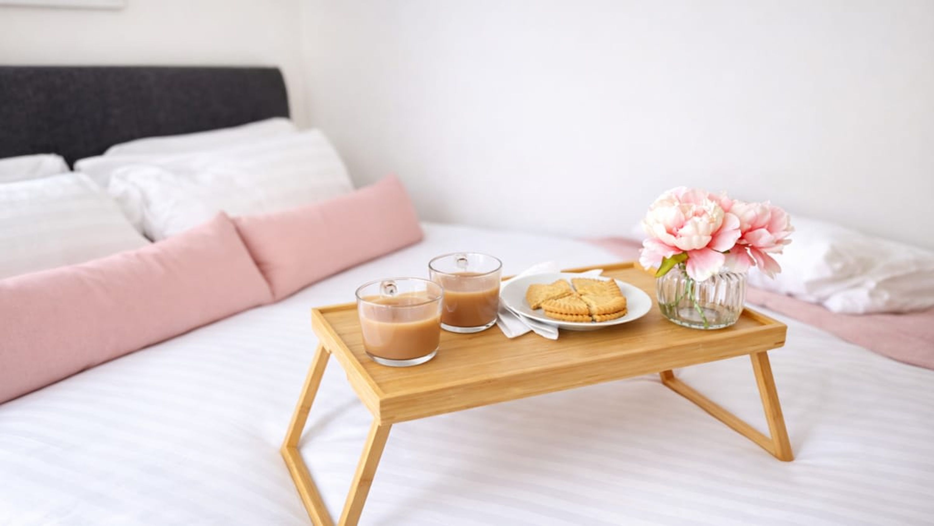 Una cama con ropa blanca, una bandeja de madera con dos vasos de una bebida, un plato con galletas y un jarrón con flores.