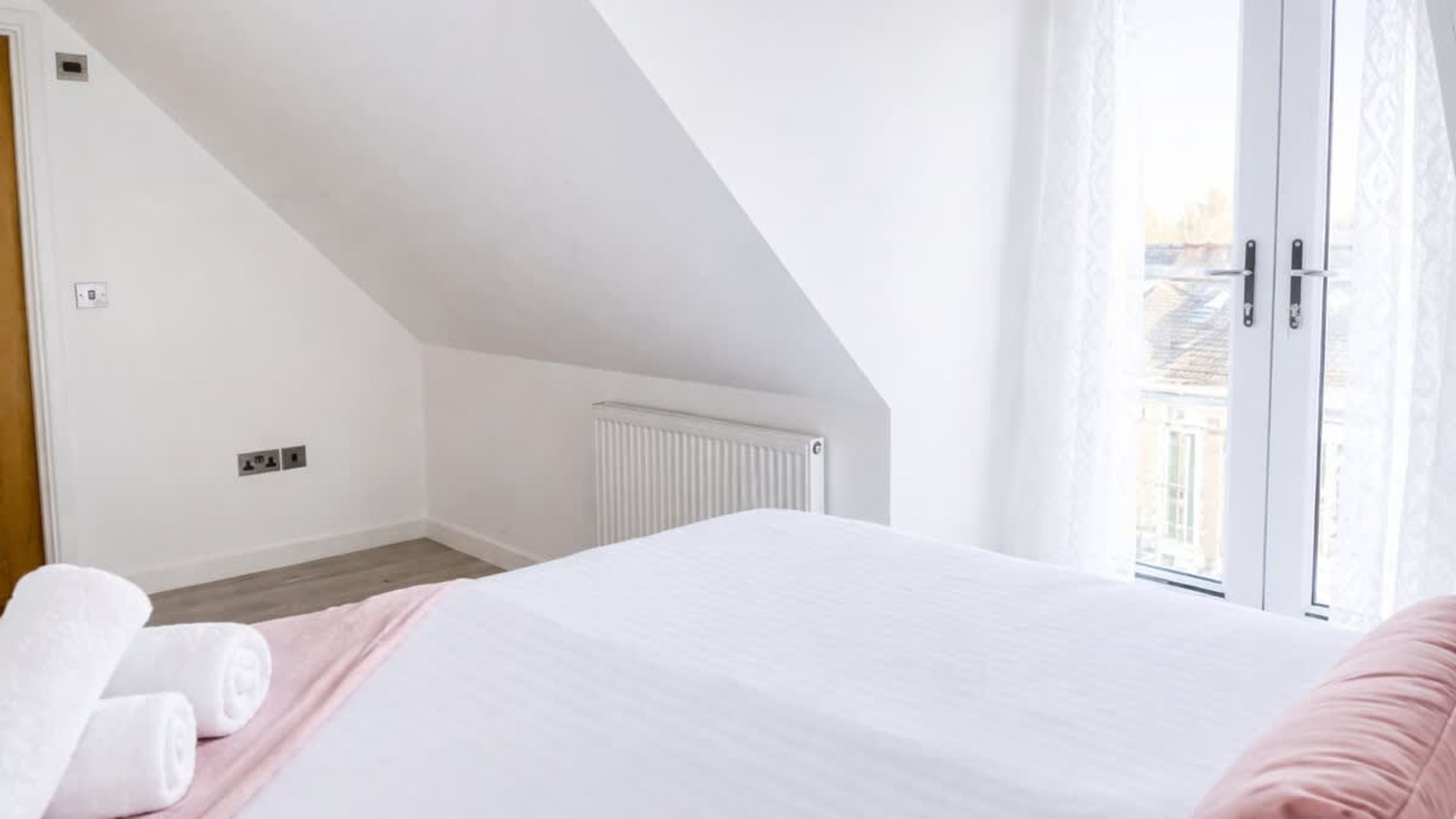 A bedroom with a sloped ceiling, a radiator, a bed with white bedding, a pink pillow, and a window with a view of buildings.