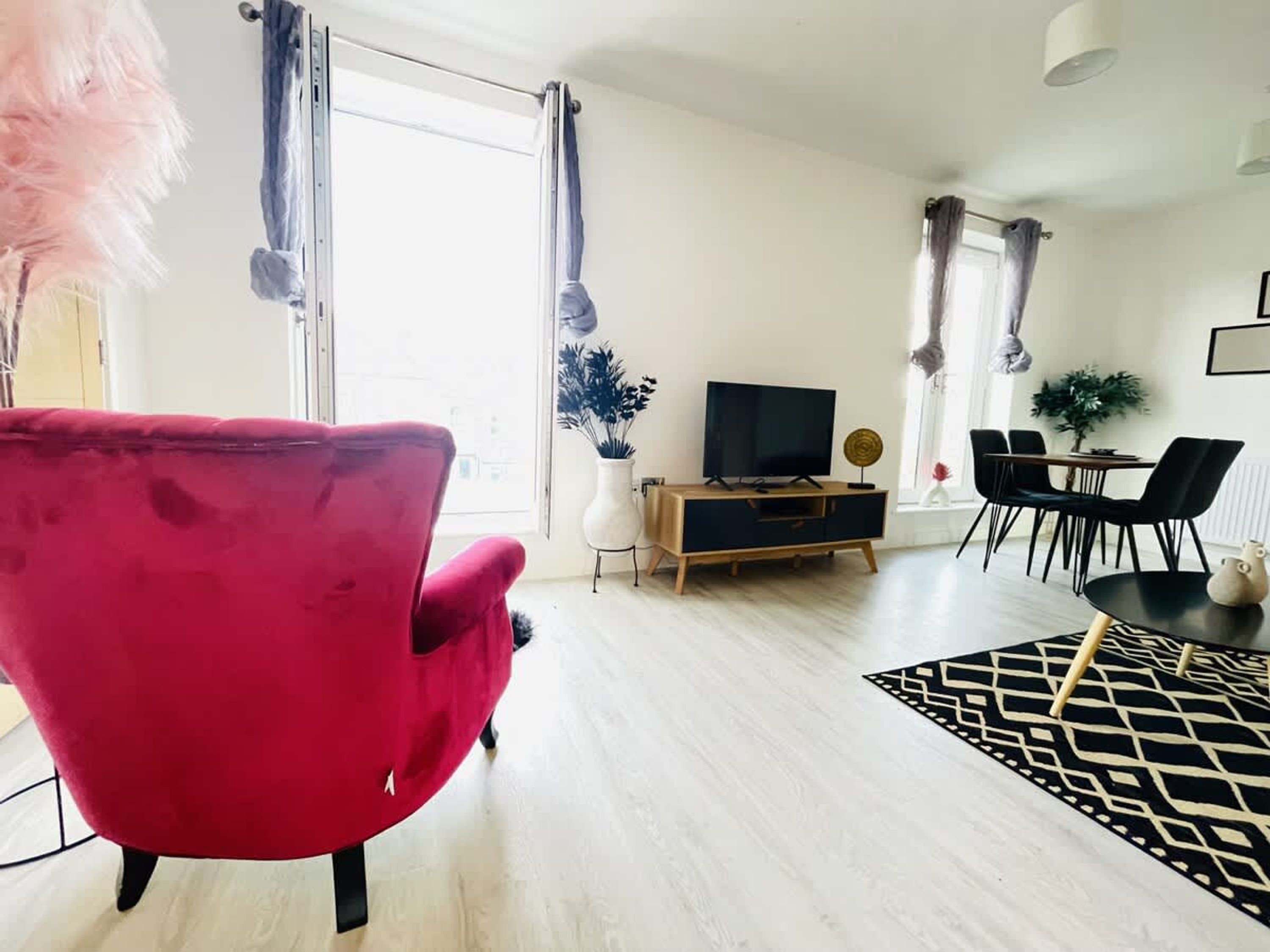 Un salon moderne avec un fauteuil rouge, un téléviseur posé sur un meuble en bois, un coin repas avec des chaises et un tapis à motifs noir et blanc.