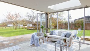 Ein großzügiger Wohnbereich mit einem großen Glasfenster, das in einen Garten führt, ein weißes Sofa, ein Couchtisch und ein kleiner Beistelltisch mit einer Lampe.