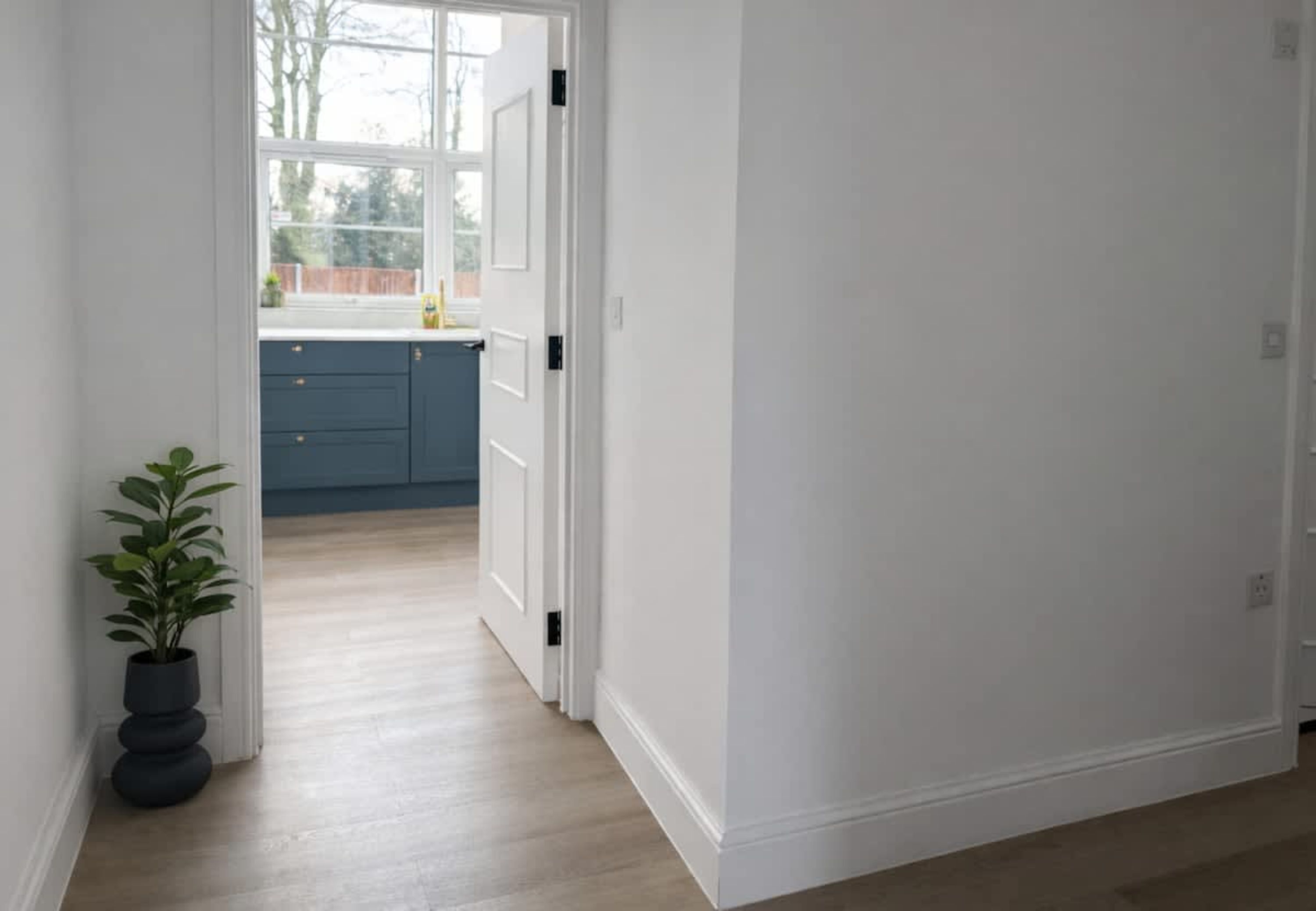 A hallway leading to a kitchen with a potted plant, wooden flooring, and a view through a window.