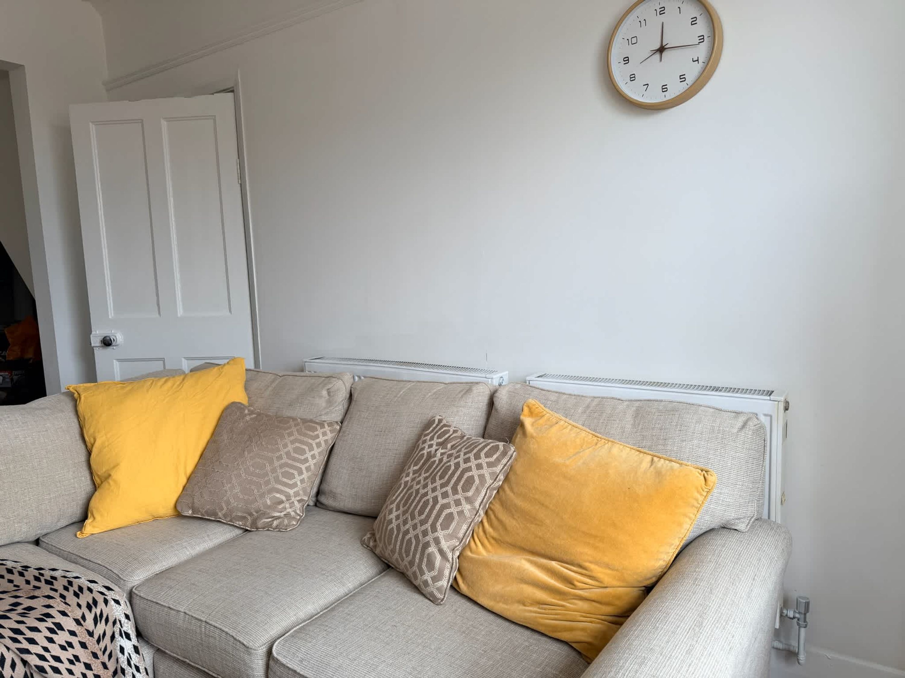 A neutral-colored sofa with patterned cushions, a wall clock, and a radiator.