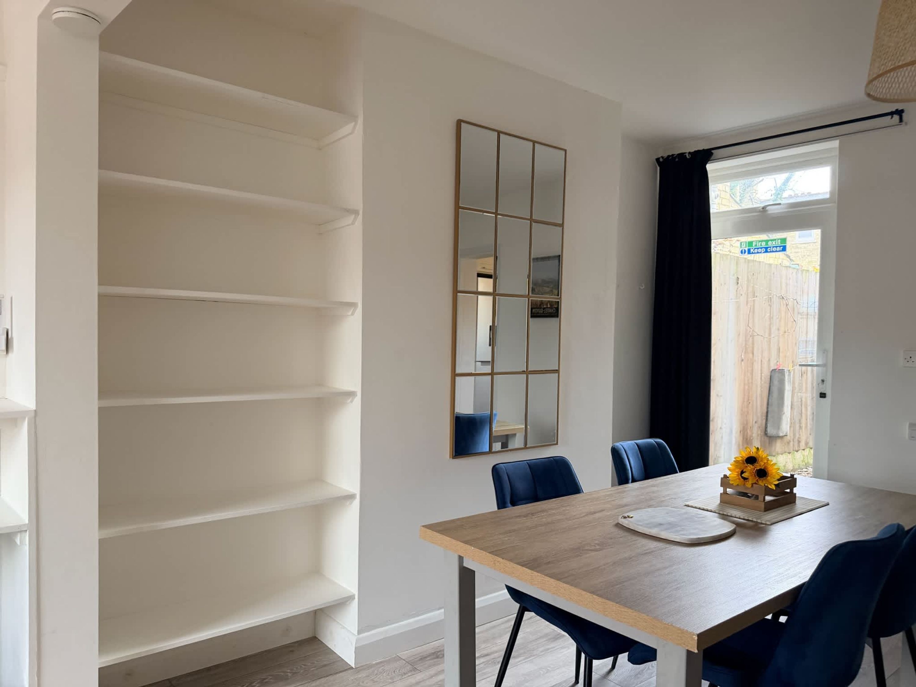 Un coin repas avec une table en bois, des chaises bleues, un miroir et une fenêtre donnant sur une clôture en bois.