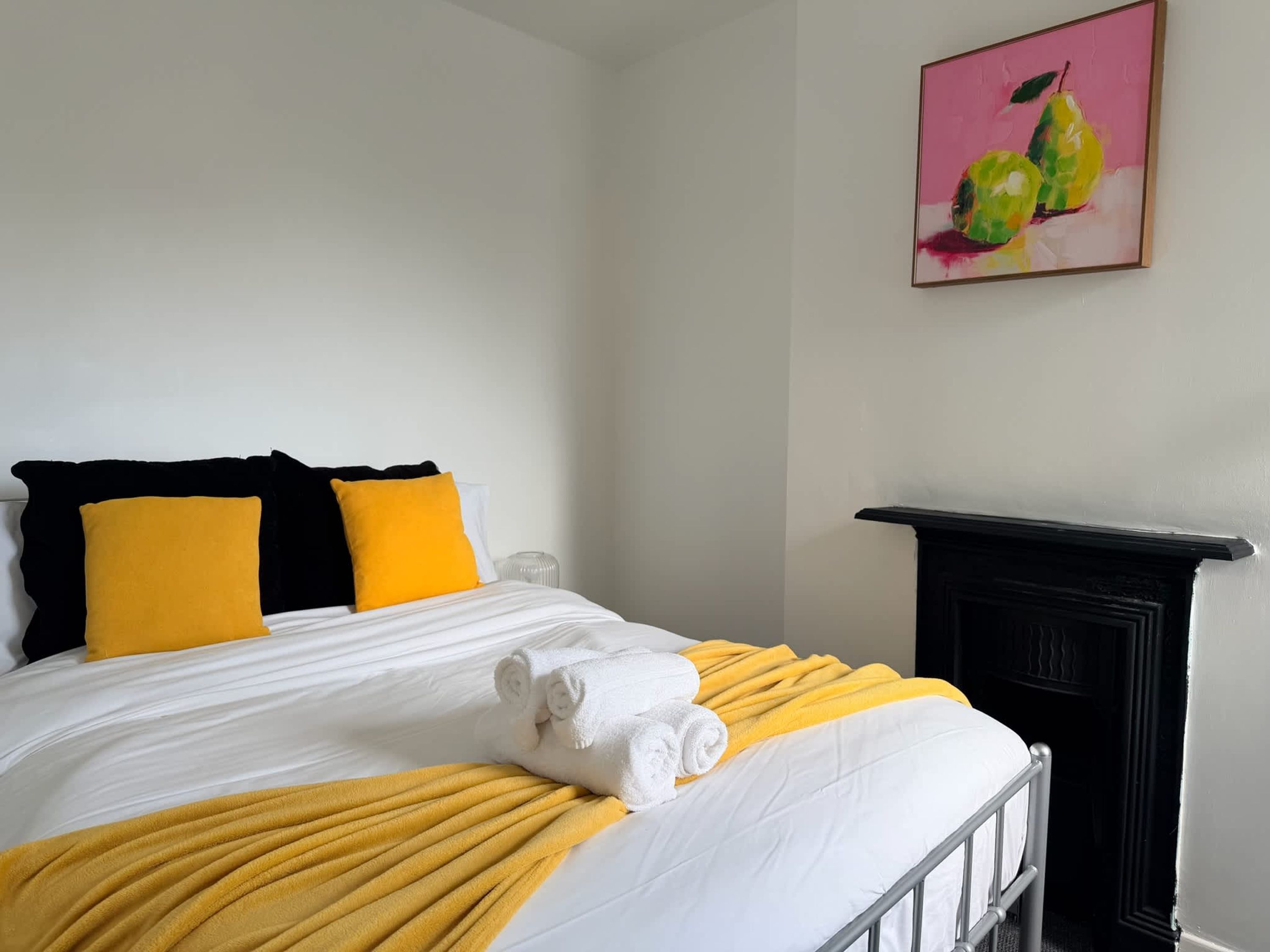 Una cama bien hecha con ropa de cama blanca y amarilla, un cabecero negro y un cuadro de peras en la pared.