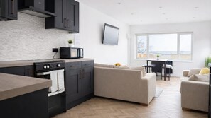 Eine moderne Küche mit dunklen Schränken, einer Mikrowelle und einem Herd. Im Wohnbereich befindet sich eine beige Sofa und ein Esstisch mit Stühlen. Ein Flachbildfernseher ist an der Wand montiert.