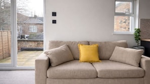 Ein beiges Sofa mit einem gelben Kissen in einem Zimmer mit großem Fenster, einer Ziegelwand und einer Pflanze.