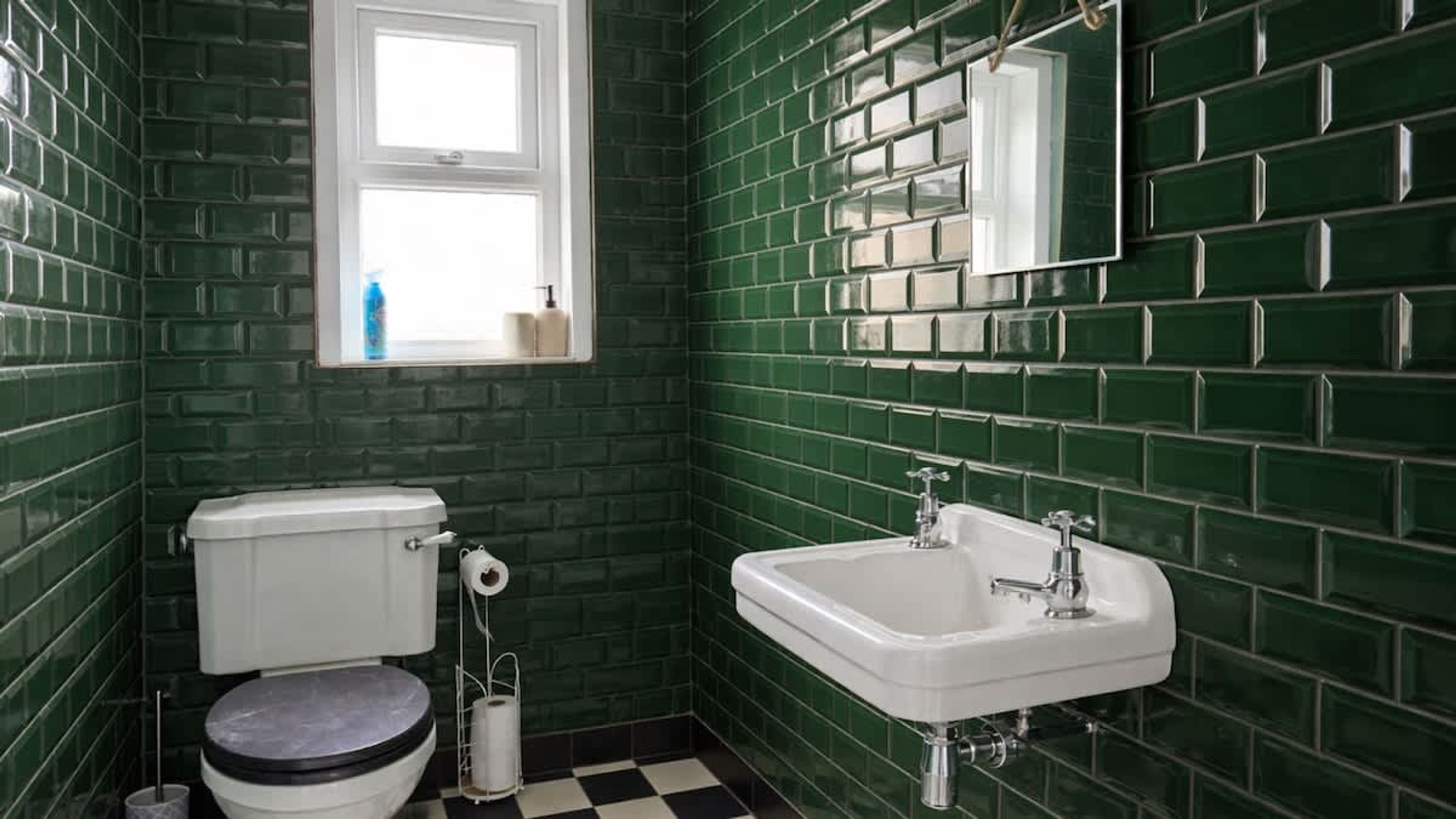 A bathroom with green tiled walls, a white toilet, a white sink, and a window above the toilet.