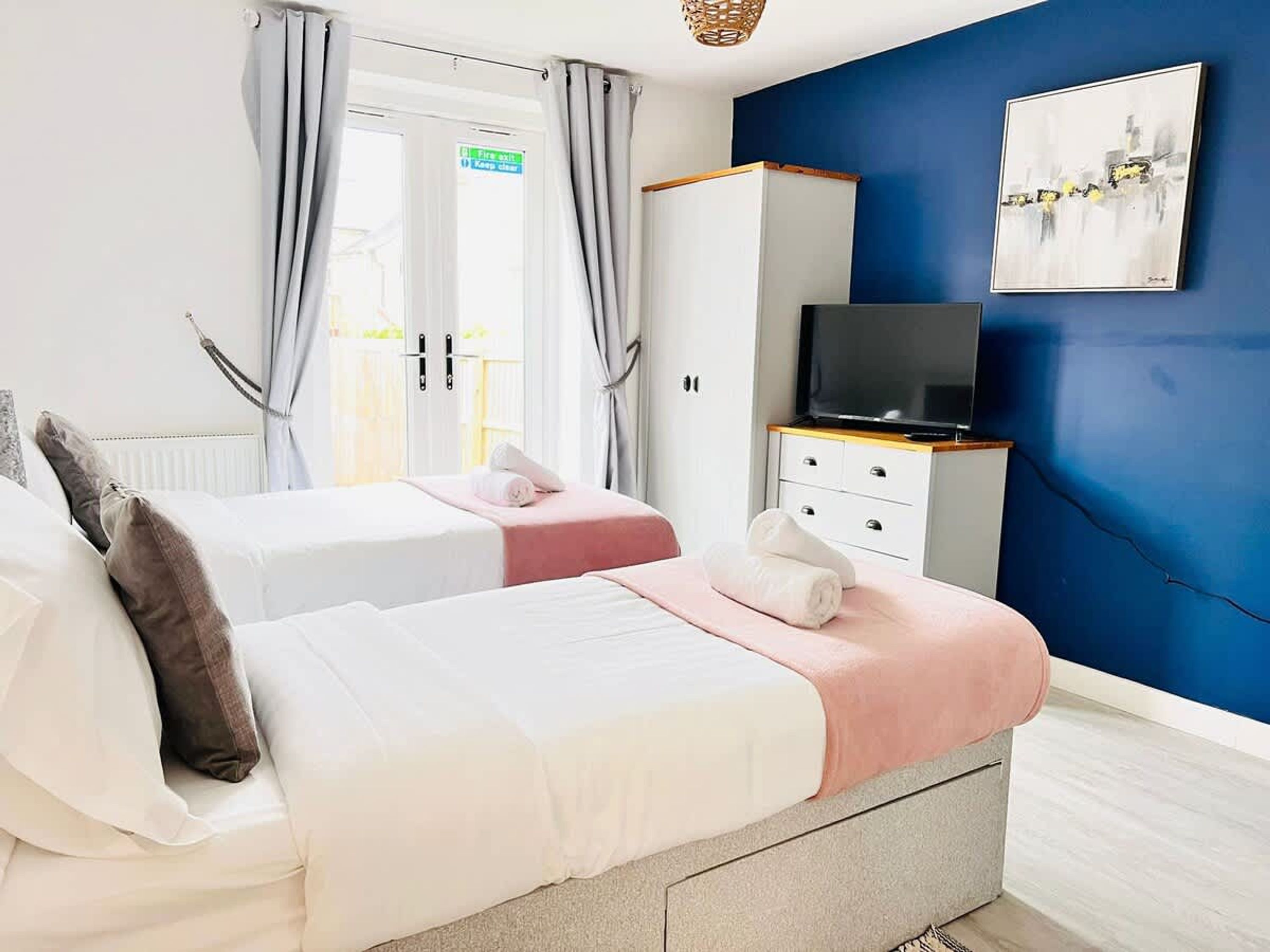 A double bed with a pink and white bedspread, a television on a wooden stand, and a blue wall.
