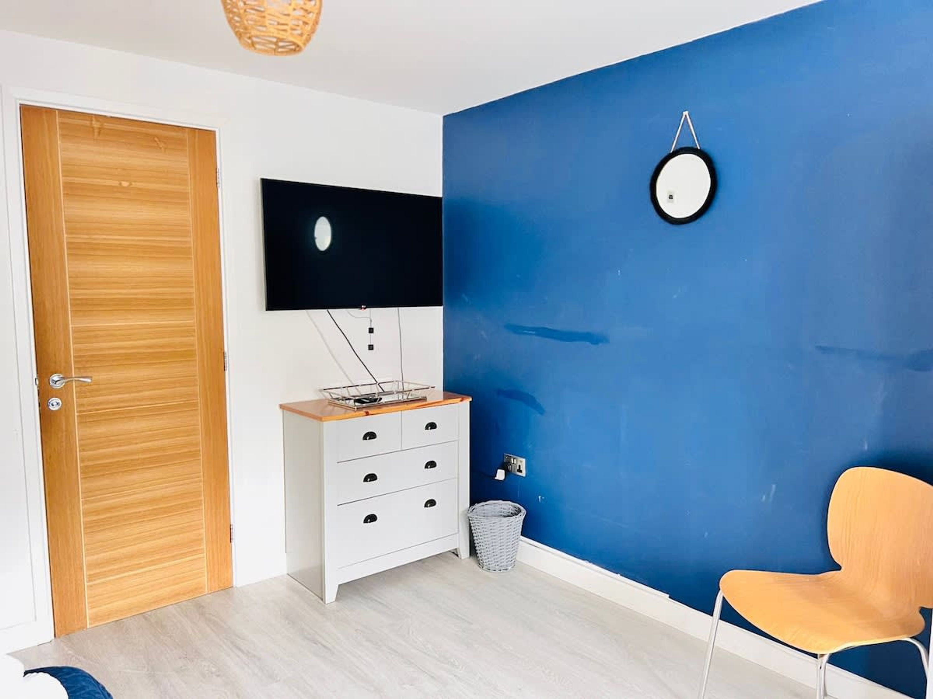 A room with a wooden door, a white dresser with drawers, a flat-screen TV mounted on the wall, a round wall clock, and a single orange chair.