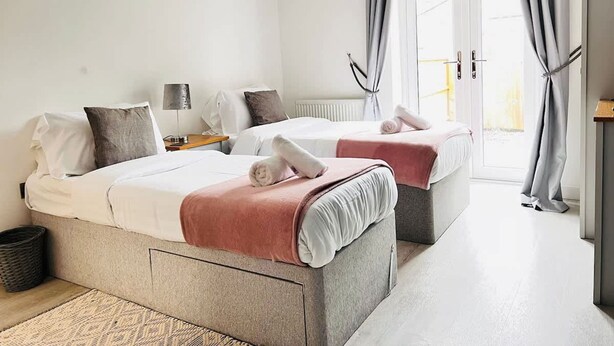A hotel room with two beds, a bedside table with a lamp, a radiator, and a view of the outside through a door. - Tanzanite Ground Floor 2BR Apart Pet Friendly (Cambridge)