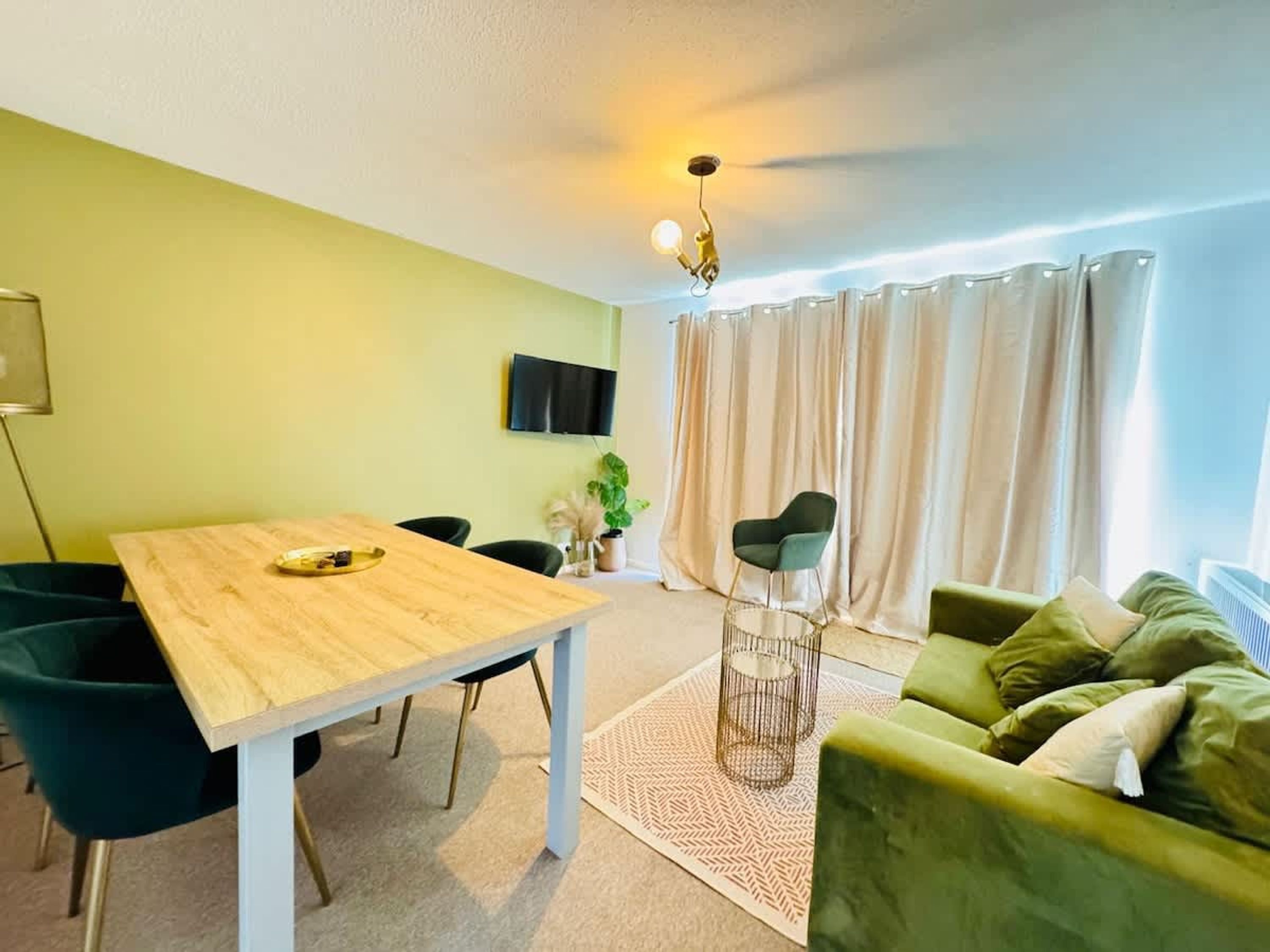 A room with a wooden table, green chairs, a green sofa, a TV on the wall, and a window with curtains.