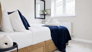 Una cama bien tendida con ropa de cama blanca y azul marino, un plaid azul oscuro y un florero blanco sobre una mesita de noche.