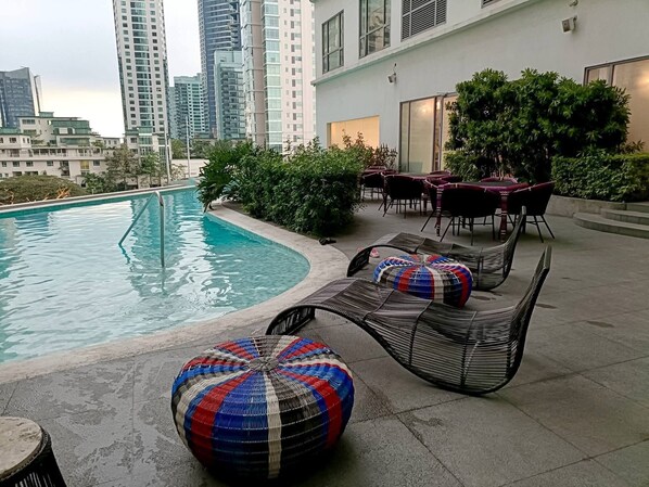 A rooftop pool area with lounge chairs and a dining table, surrounded by greenery and overlooking a cityscape. - Room-lerfo Staycation!!!! (Mandaluyong)