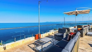Eine Dachterrasse mit Sitzgelegenheiten im Freien, einer Glasbrüstung und Blick aufs Meer.