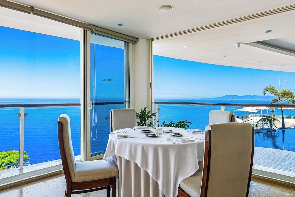 Un comedor con una mesa preparada para cuatro, ventanas amplias con vista al mar y una piscina visible al fondo.