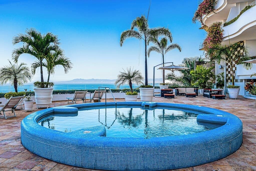 A circular pool with lounge chairs and potted plants, overlooking a clear blue sea.