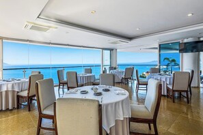 A restaurant with round tables, chairs, and a view of the sea. - Elegant Suite With Ocean-view Pools, hot Tub, Gym, Balcony & Full Kitchen (Puerto Vallarta)