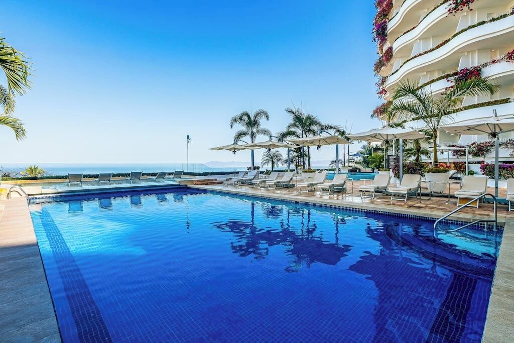 Ein Pool mit Liegestühlen und Sonnenschirmen, umgeben von Palmen und Blick auf den Ozean.