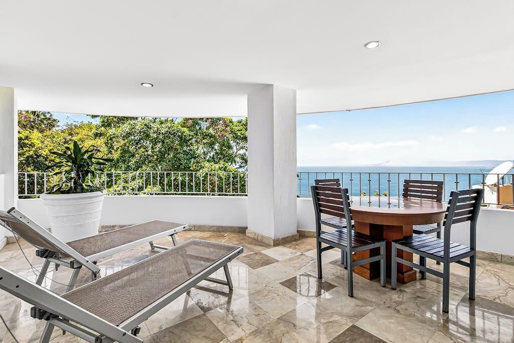 Een balkon met uitzicht op zee, een ronde tafel met stoelen en twee ligbedden.