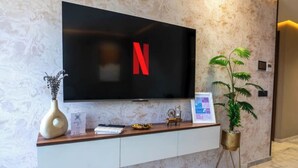 Ein modernes Wohnzimmer mit einem Flachbildfernseher, der das Netflix-Logo zeigt, einem weißen Konsolentisch mit Dekoration und einer Topfpflanze.