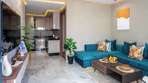 Ein modernes Wohnzimmer mit einem türkisfarbenen Sofa, einem Couchtisch mit Obst, einer Topfpflanze und einer Küche im Hintergrund.