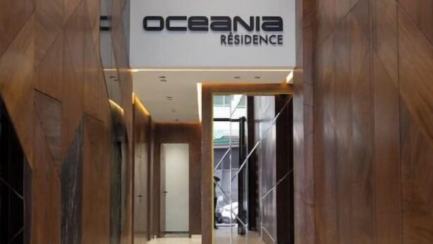 Ein Flur mit einem Schild für 'Oceania Residence', der zu einem Raum mit einem großen Spiegel und einer blauen Mülltonne führt.