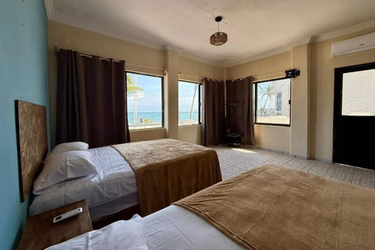 A hotel room with two beds, a window with curtains, a ceiling fan, and a view of the ocean.