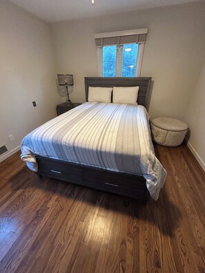 Ein Schlafzimmer mit Bett, Nachttisch, Lampe, Fenster und einem runden Hocker.