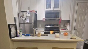 Cocina con armarios blancos, un refrigerador de acero inoxidable, un microondas y un lavamanos. Hay dos taburetes de madera frente a la encimera.
