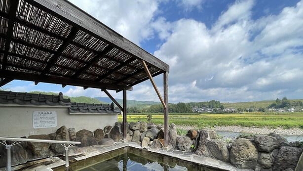 A stone bath area with a wooden roof, a stone wall, and a view of a green landscape. - BungonoSato (Kusu)