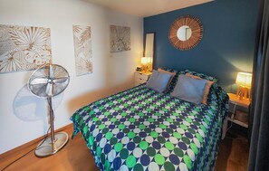 A bedroom with a patterned bedspread, a standing fan, a mirror, and wall art. - Appartment proche plage (Valras Plage)