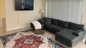 A modern living room with a sectional sofa, a flat-screen TV, a patterned rug, and a window with blinds. - Beautiful 3-bedroom house with AC in adorable New Port Richey (New Port Richey)
