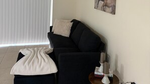 A living room with a black sofa, a white blanket, a small table with a lamp, and a painting on the wall. - Beautiful 3-bedroom house with AC in adorable New Port Richey (New Port Richey)