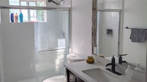 A modern bathroom with a glass shower enclosure, a white toilet, a vanity with a mirror, and a window providing natural light.