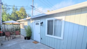 A single-story building with a front door, a window, and a small patio area with chairs and a potted plant.