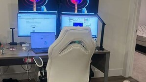 Ein Zimmer mit Schreibtisch, zwei Monitoren, einem Laptop, einem weißen Bürostuhl und einem Bett.