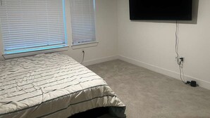 Ein Schlafzimmer mit einem Bett, zwei Fenstern mit Jalousien, einem an der Wand befestigten Fernseher und einem Teppichboden.