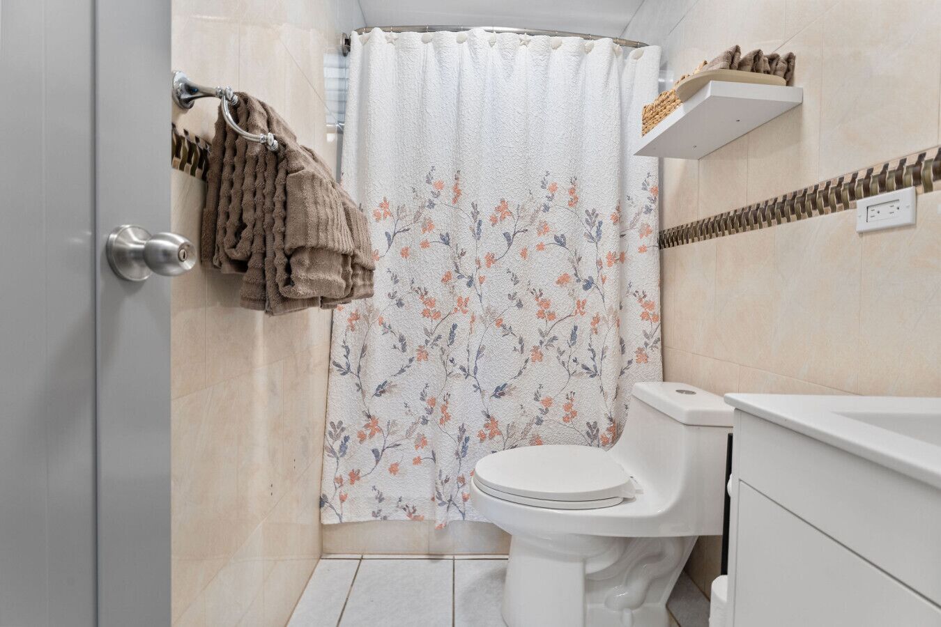 Un baño con inodoro, cortina de ducha estampada con motivos florales, toallero con toallas y repisa con artículos de aseo.