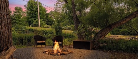 A fire pit with two chairs and a wooden bench in a natural setting.