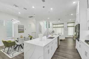 Cozinha moderna com ilha de mármore, armários brancos e área de refeições com mesa e cadeiras de vidro.
