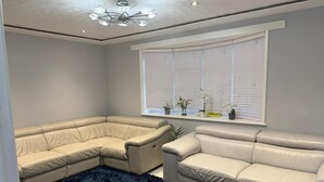A corner room with a sectional sofa, a dark blue rug, a window with blinds, and a ceiling light fixture. - Two bedroom independent house with parking & WiFi (Romford)