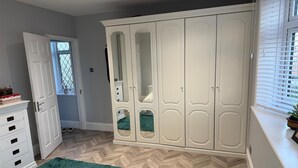 A bedroom with a large wardrobe, a bed with a floral bedspread, a green rug, and a window with blinds. - Two bedroom independent house with parking & WiFi (Romford)