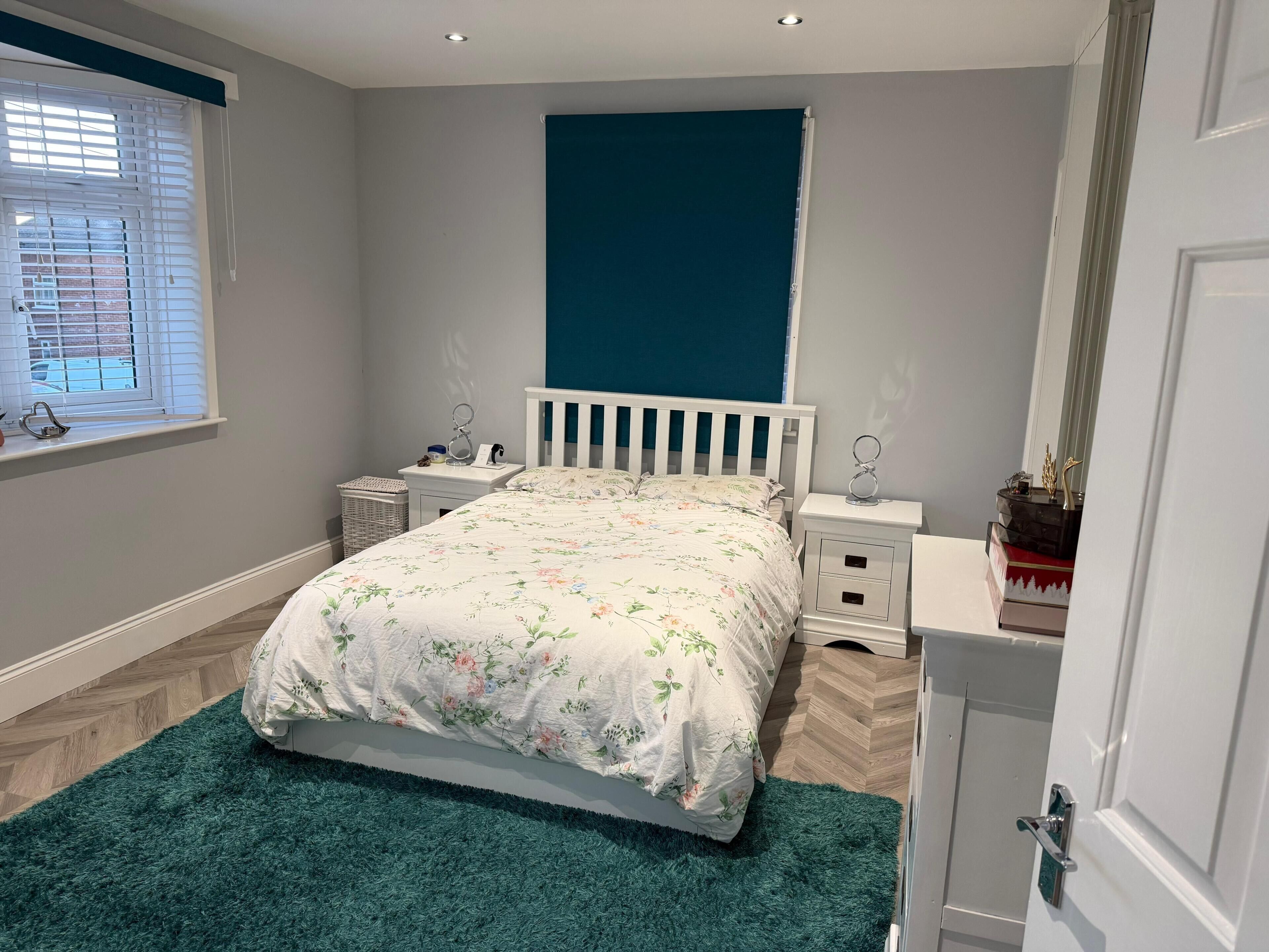 A bedroom with a bed, nightstands, a dresser, a window with blinds, and a blue accent wall.