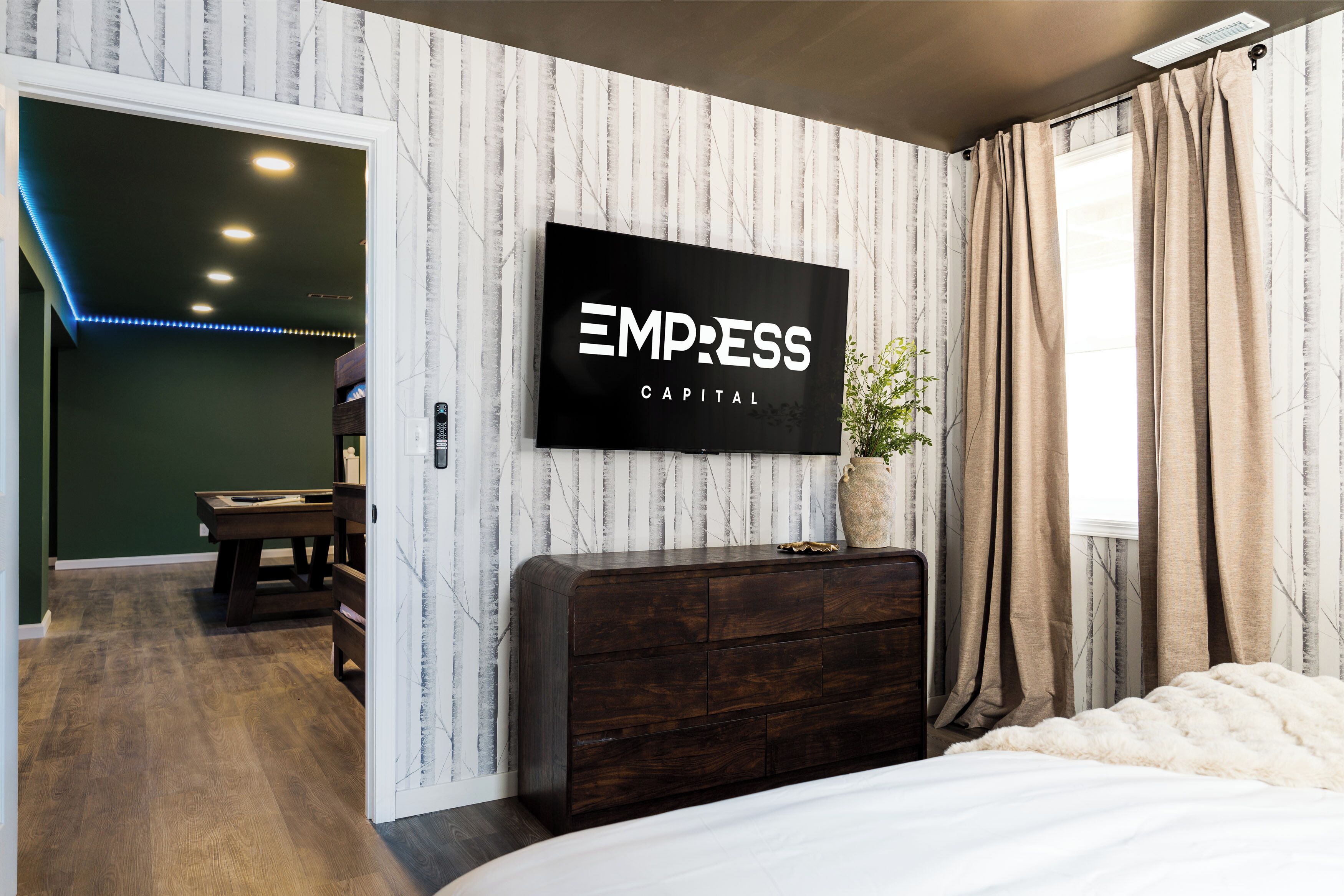 A hotel room with a dark wooden dresser, a flat-screen TV mounted on the wall, a window with curtains, and a plant on the dresser.