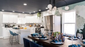 Eine moderne Küche mit weißen Schränken, einem Edelstahl-Kühlschrank und einem Essbereich, gedeckt für sechs Personen, mit blauen Servietten und Kerzen.