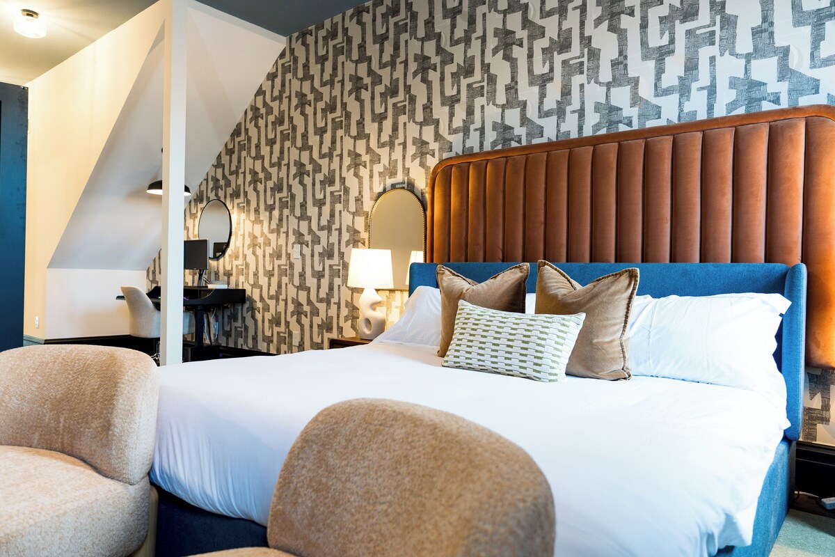 A hotel room with a large bed, a headboard, a desk with a lamp, and a patterned wall.