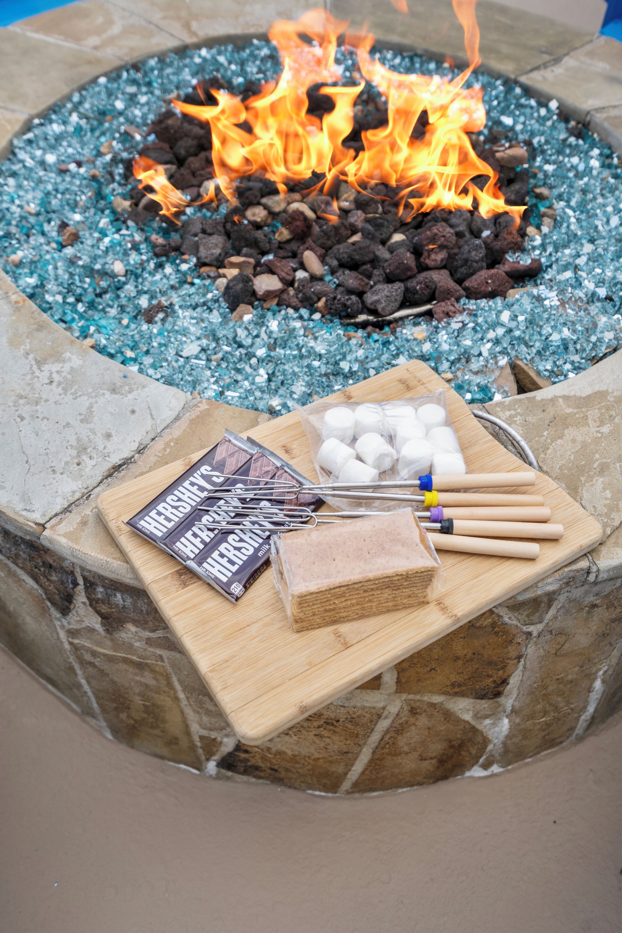 A stone fire pit with a fire burning, surrounded by blue glass pebbles. In front of the fire, there is a wooden board with marshmallows, chocolate bars, and a candle.